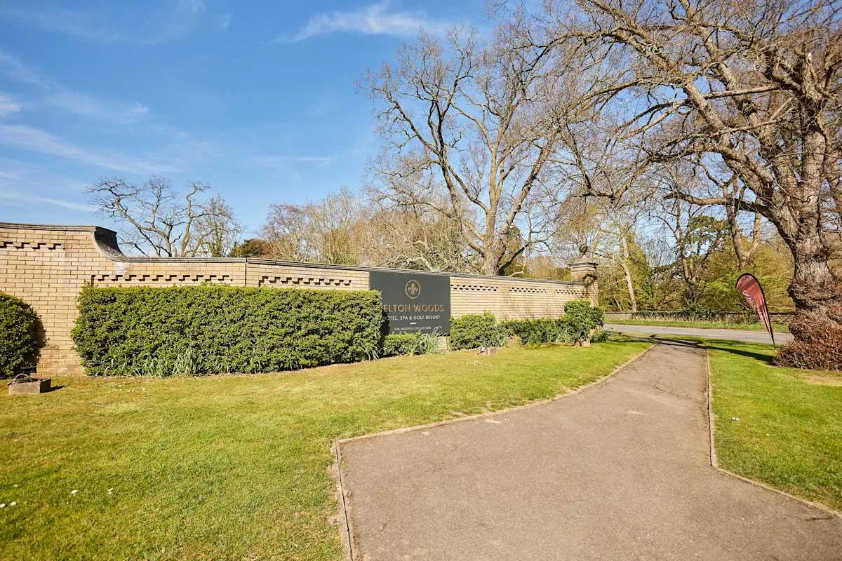 A paved path curves through a grassy entrance beside a brick wall and trees.