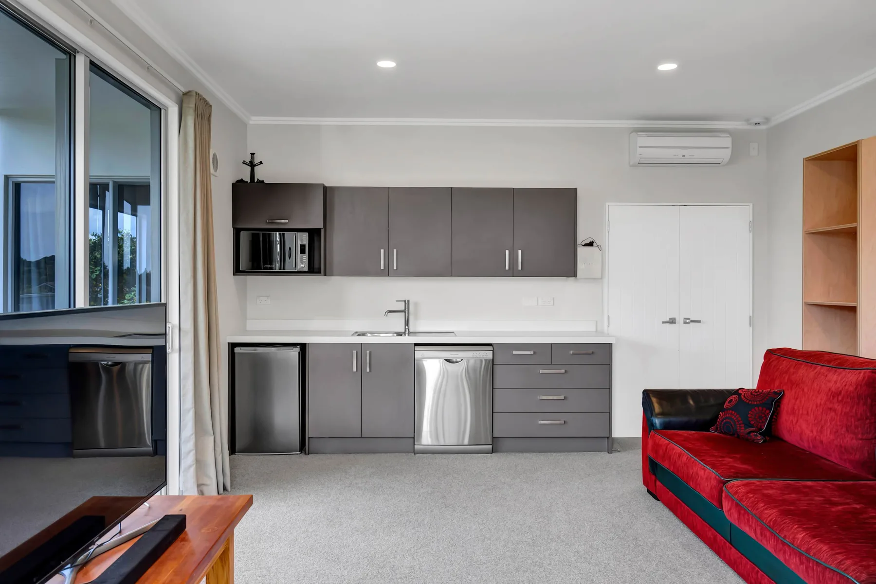 Una sala de estar con cocina compacta y asientos rojos junto a grandes ventanas.