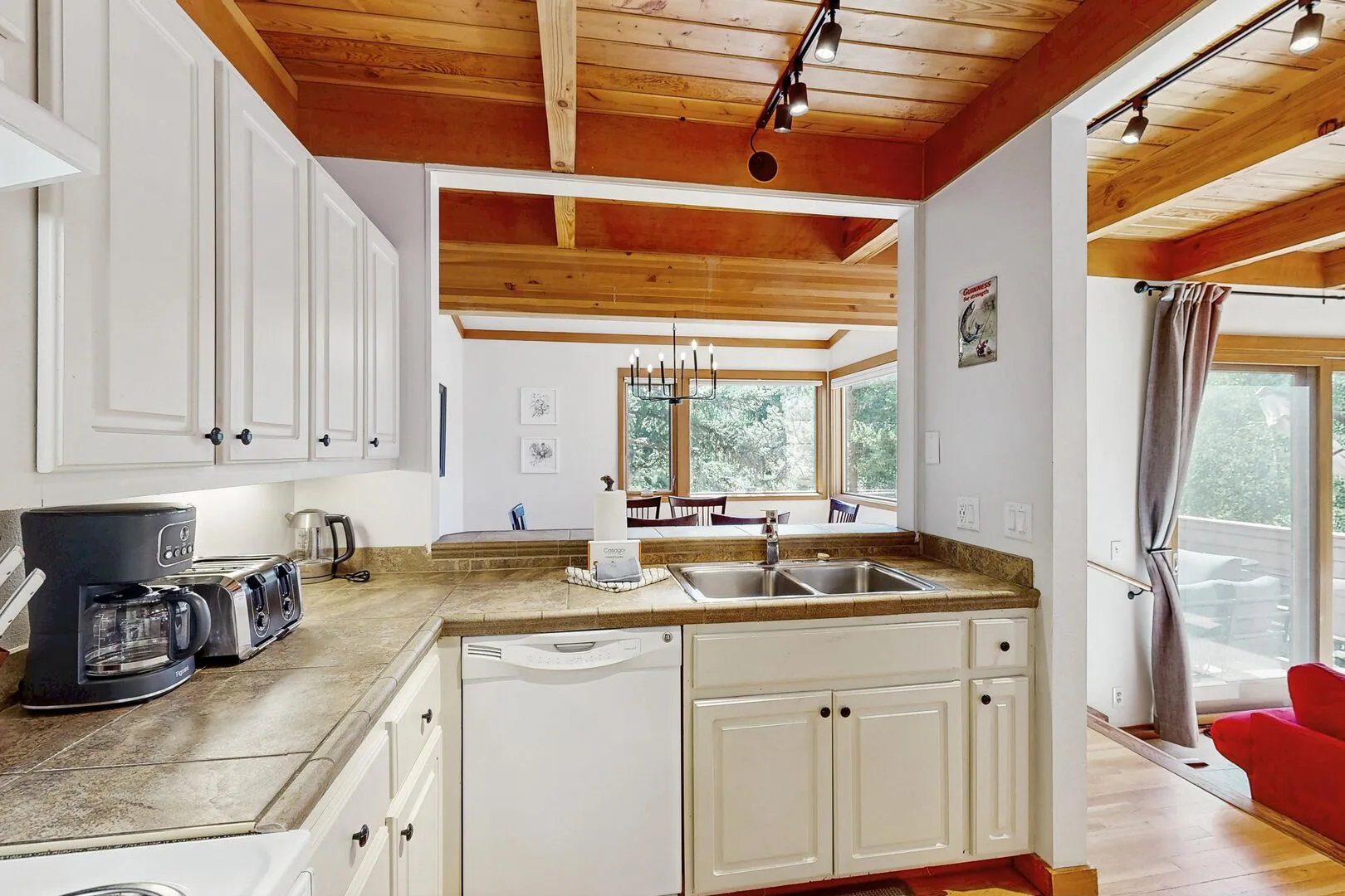 A bright kitchen with white cabinets, wood ceiling beams, and a counter overlooking a window.