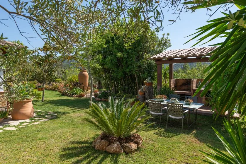 Un patio de jardín con asientos al aire libre bajo una pérgola de tejas, rodeado de vegetación.