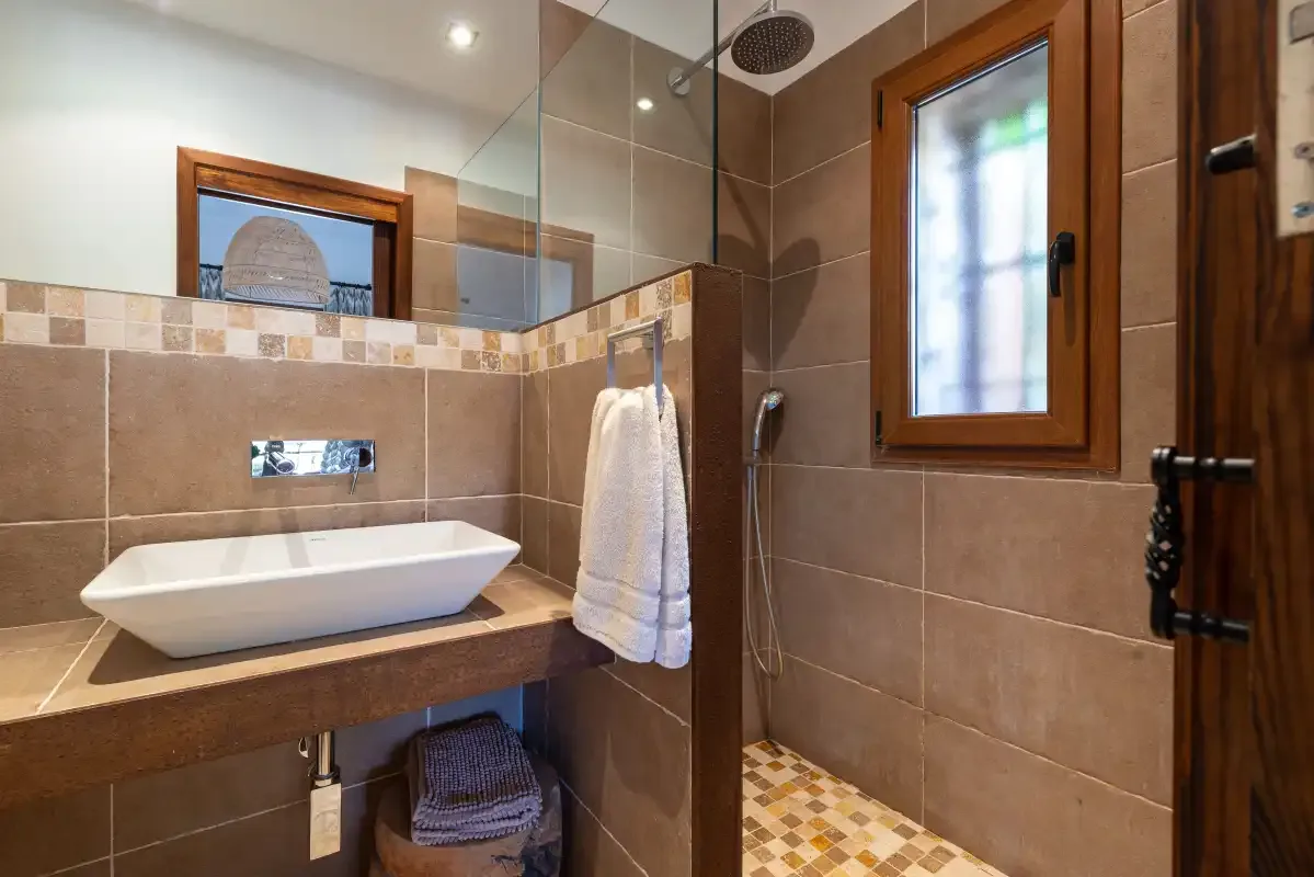 A tiled bathroom with a sink, shower, and wooden-framed window.