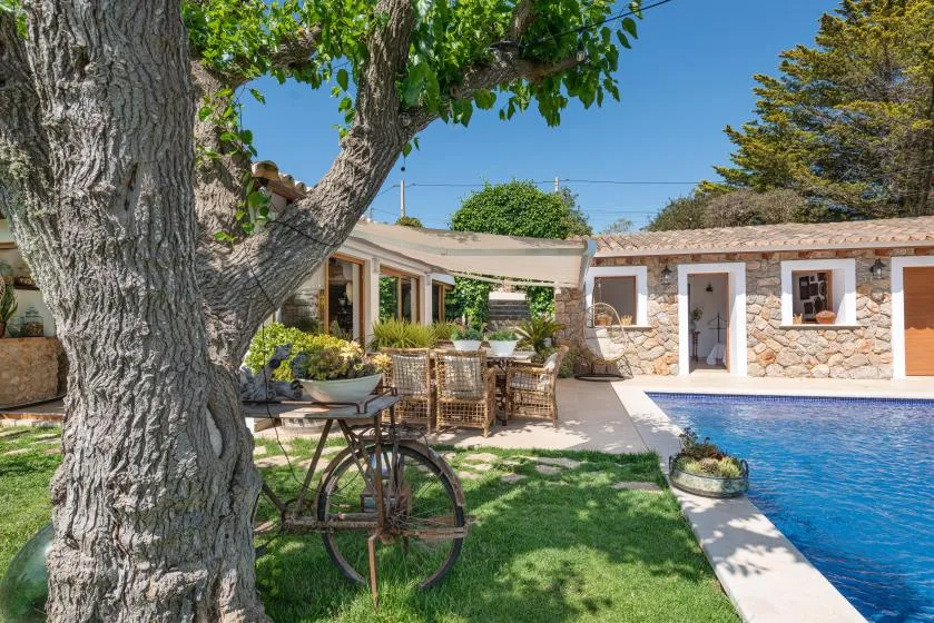 A sunny outdoor patio with seating beside a swimming pool and leafy trees.