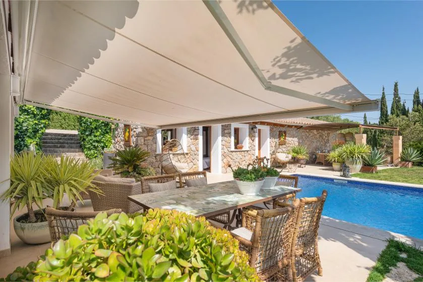 A shaded outdoor dining area beside a swimming pool, surrounded by plants.