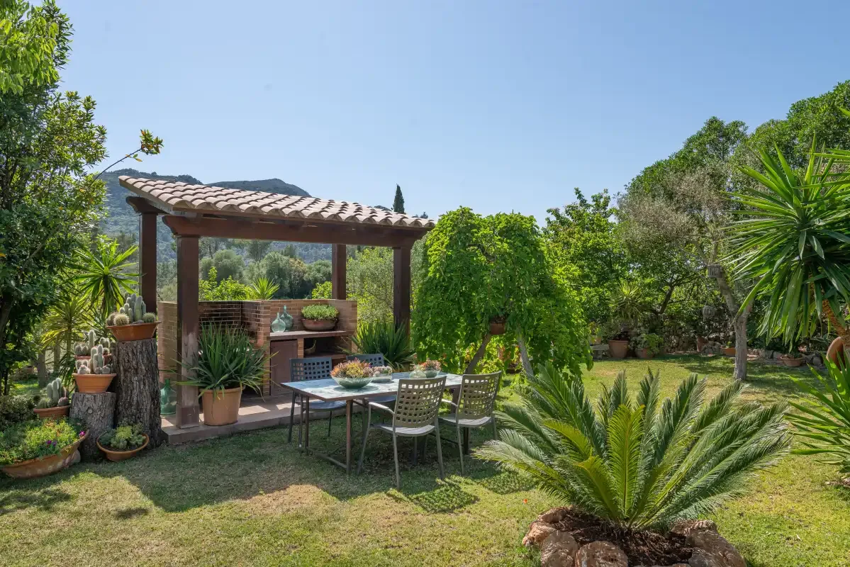 A garden patio has outdoor seating beneath a pergola, surrounded by greenery.