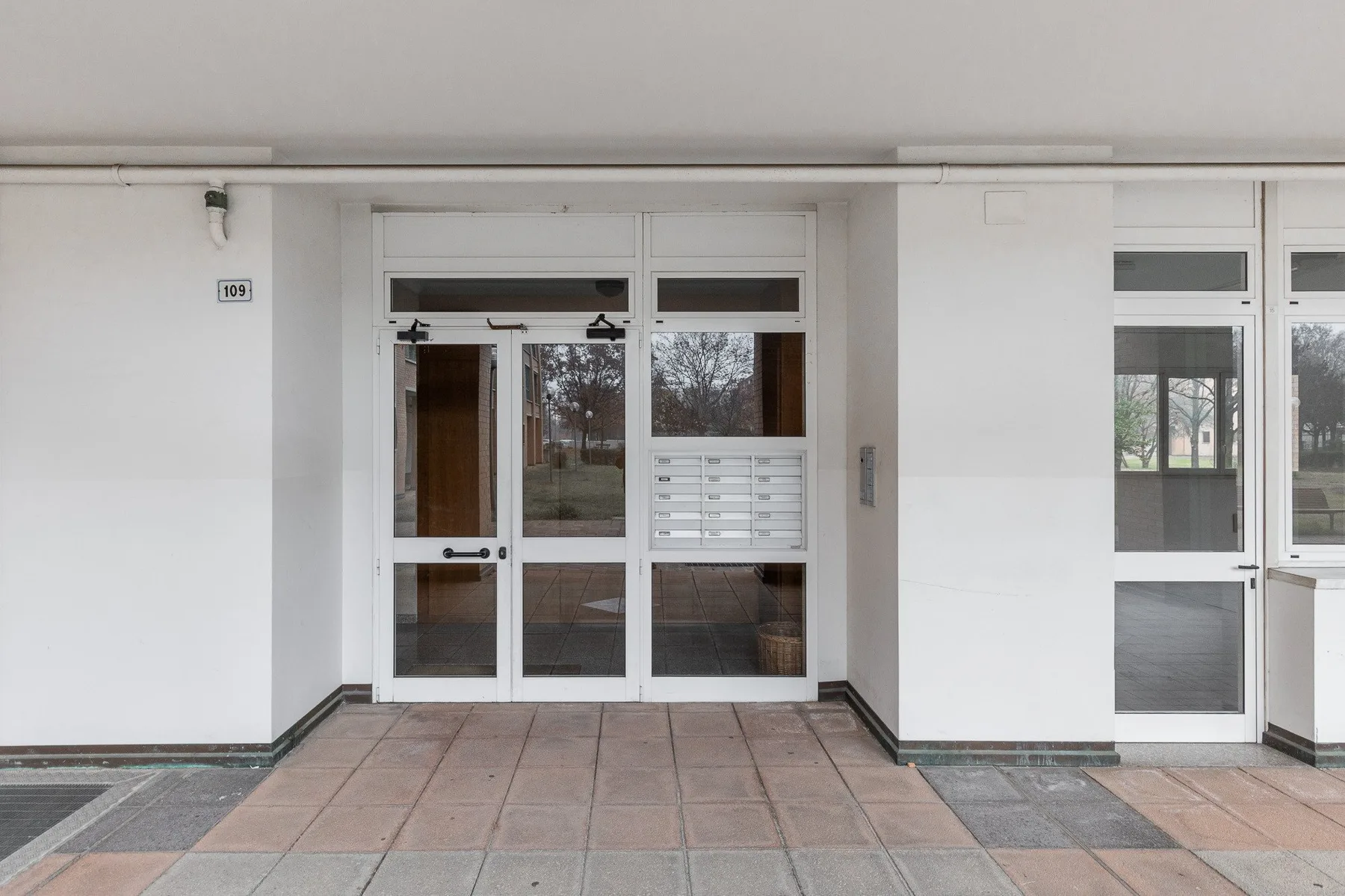 La entrada de un edificio blanco con puertas de vidrio da a un pasillo embaldosado.