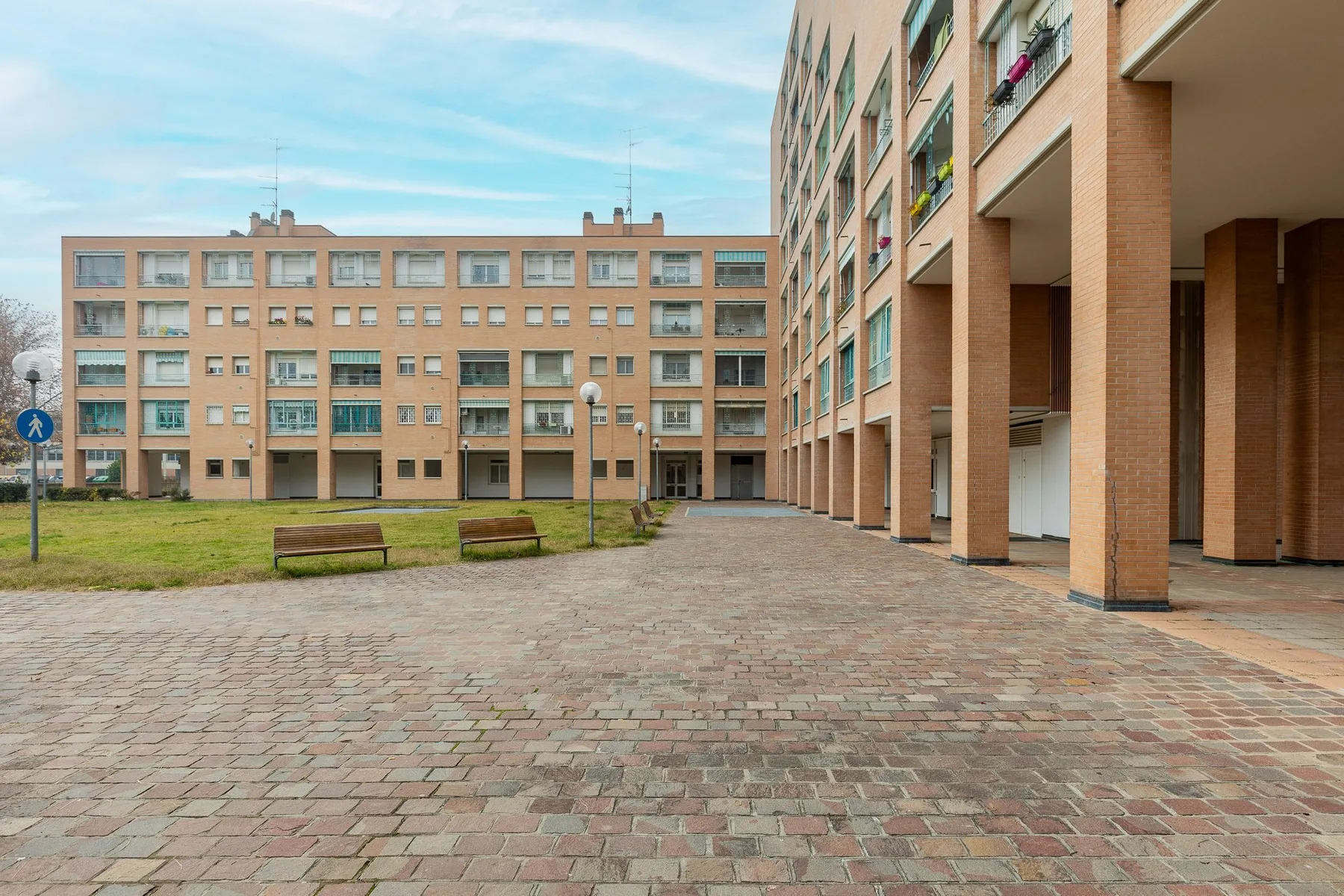 Un patio pavimentado entre edificios de ladrillo, con bancos y una zona de césped.