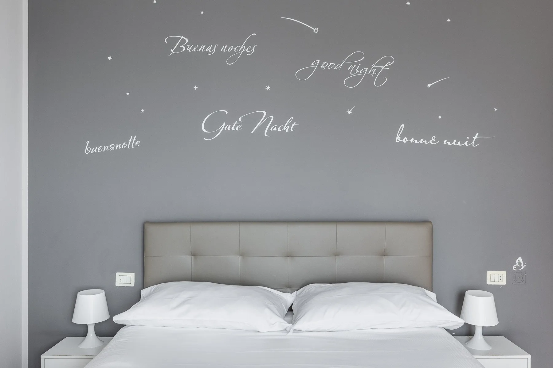 Un dormitorio con una cama tendida frente a una pared gris decorada con saludos en varios idiomas.