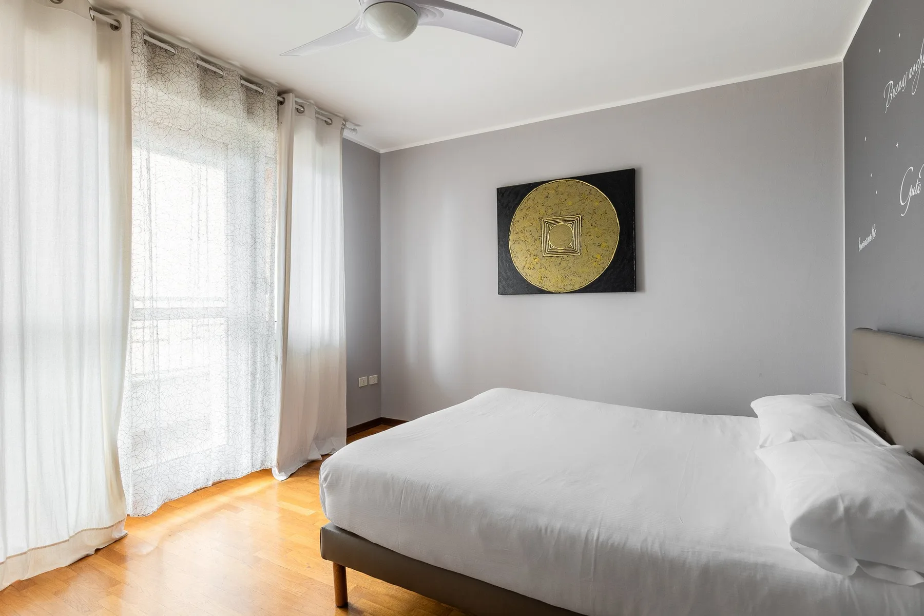 Un dormitorio con una cama, cortinas translúcidas y arte enmarcado en la pared.