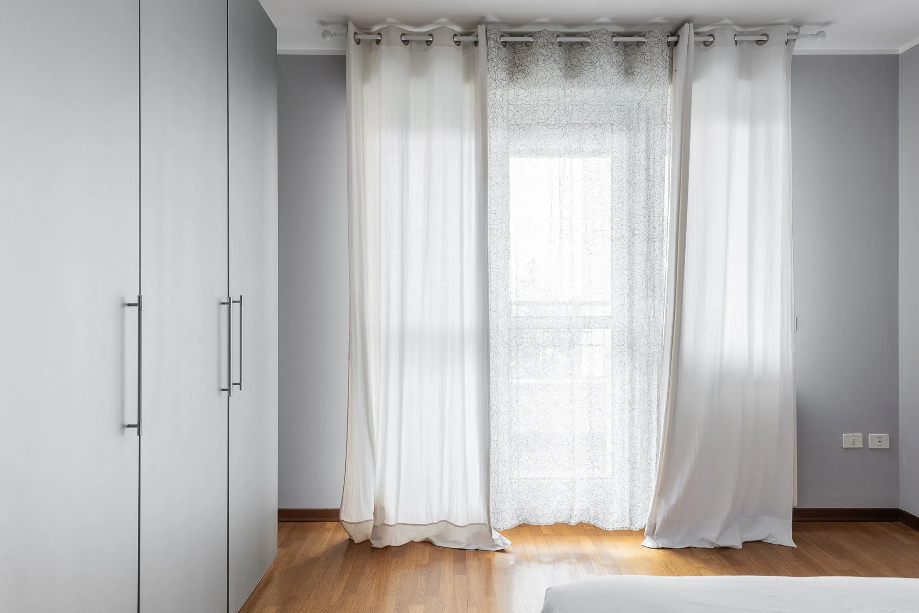 Un dormitorio luminoso con cortinas blancas en una ventana amplia y suelo de madera.
