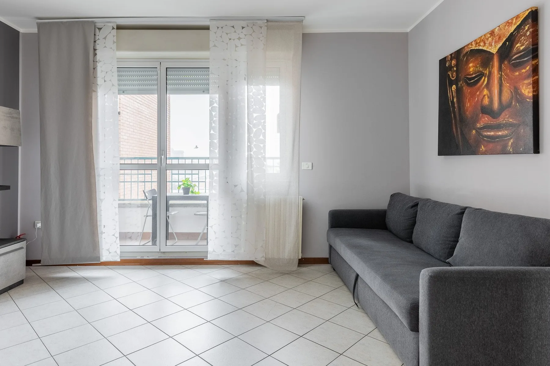 Una sala de estar con un sofá gris, suelo de baldosas y ventanas grandes.