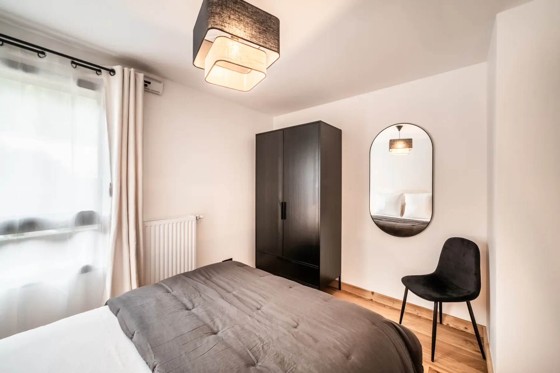 A bedroom with a gray-covered bed, a dark wardrobe, and a chair beside a mirror.