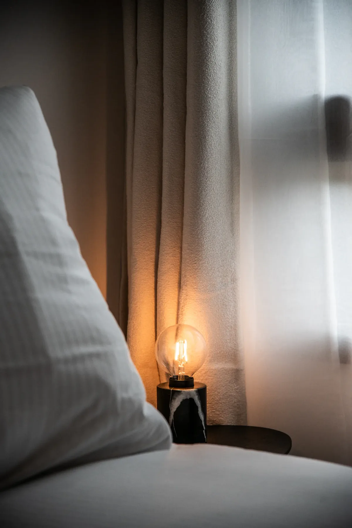 A dimly lit bedside lamp beside a white bed, with light filtering through sheer curtains in the background.