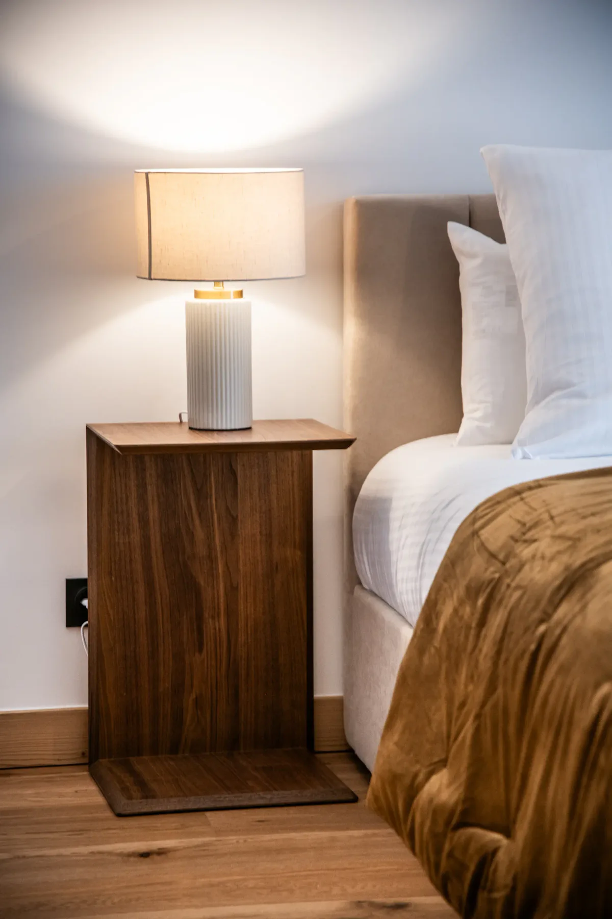 A bedroom scene with a bedside table and lamp beside a bed with white pillows and a brown blanket.
