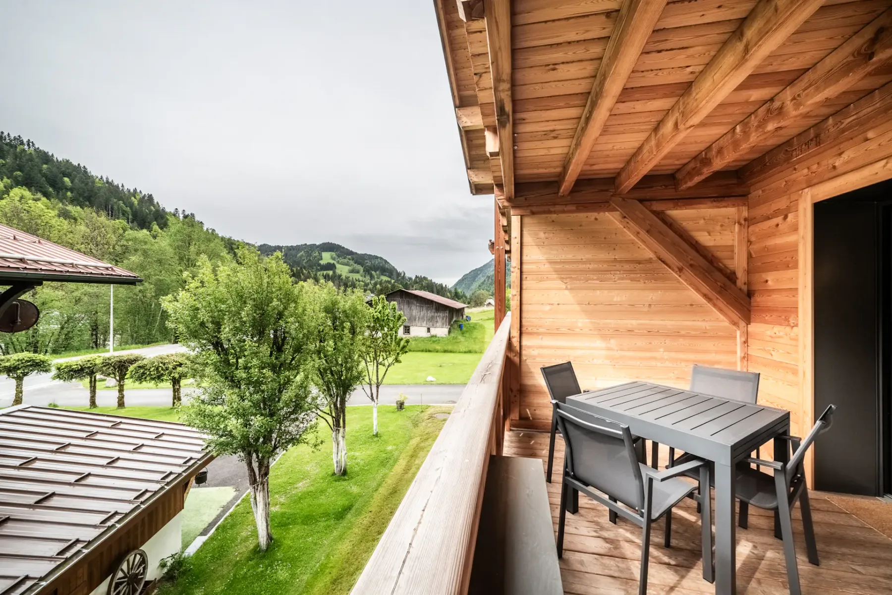 Outdoor deck or terrace with wooden roof, modern black table and chairs, and cushioned seating area. Overlooks wooded hillside and rustic buildings.
