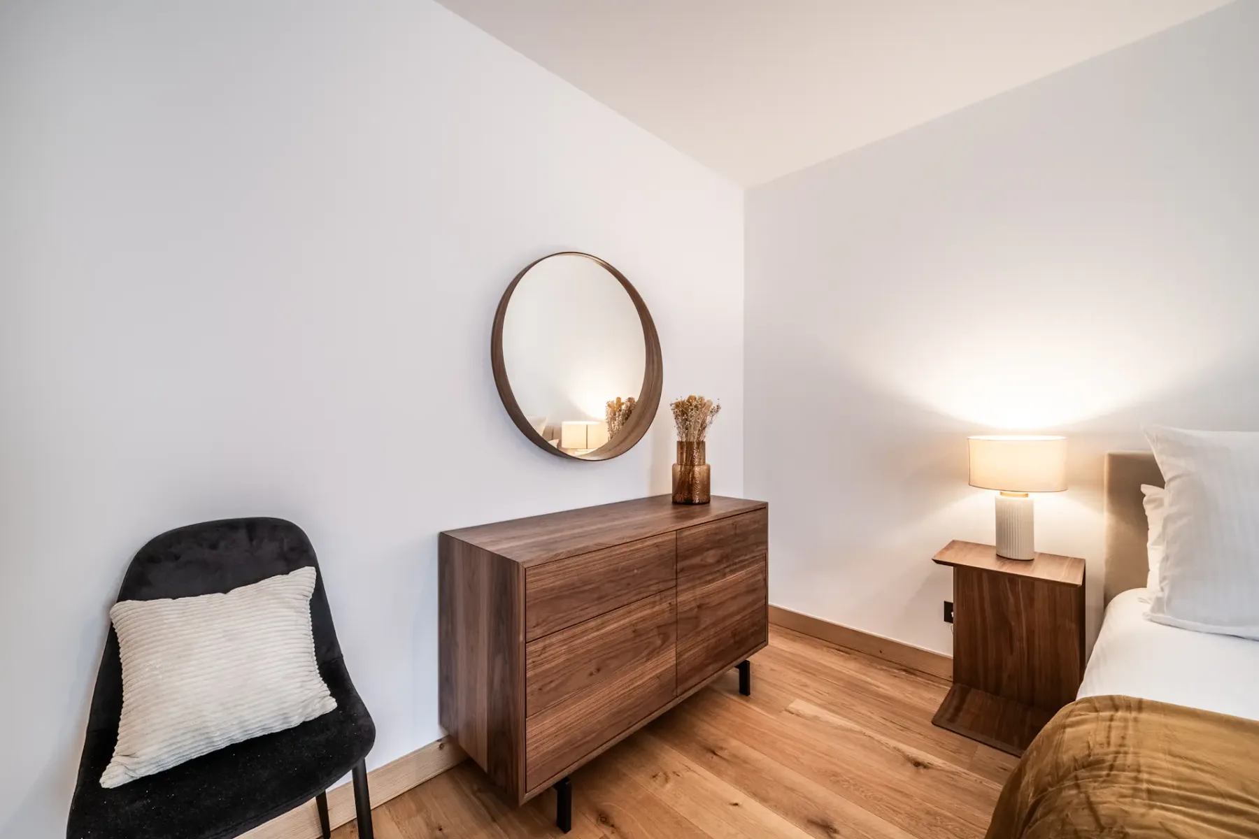 A bedroom with a bed, bedside table, and wooden dresser beneath a round mirror.