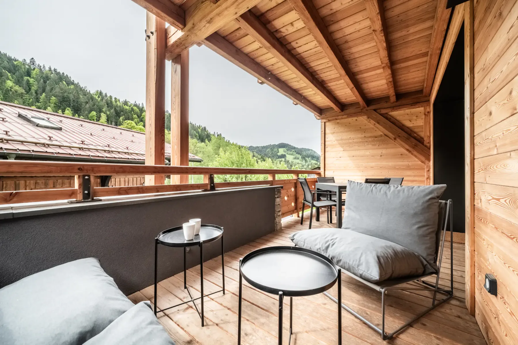Outdoor deck or terrace with wooden roof, modern black table and chairs, and cushioned seating area. Overlooks wooded hillside and rustic buildings.