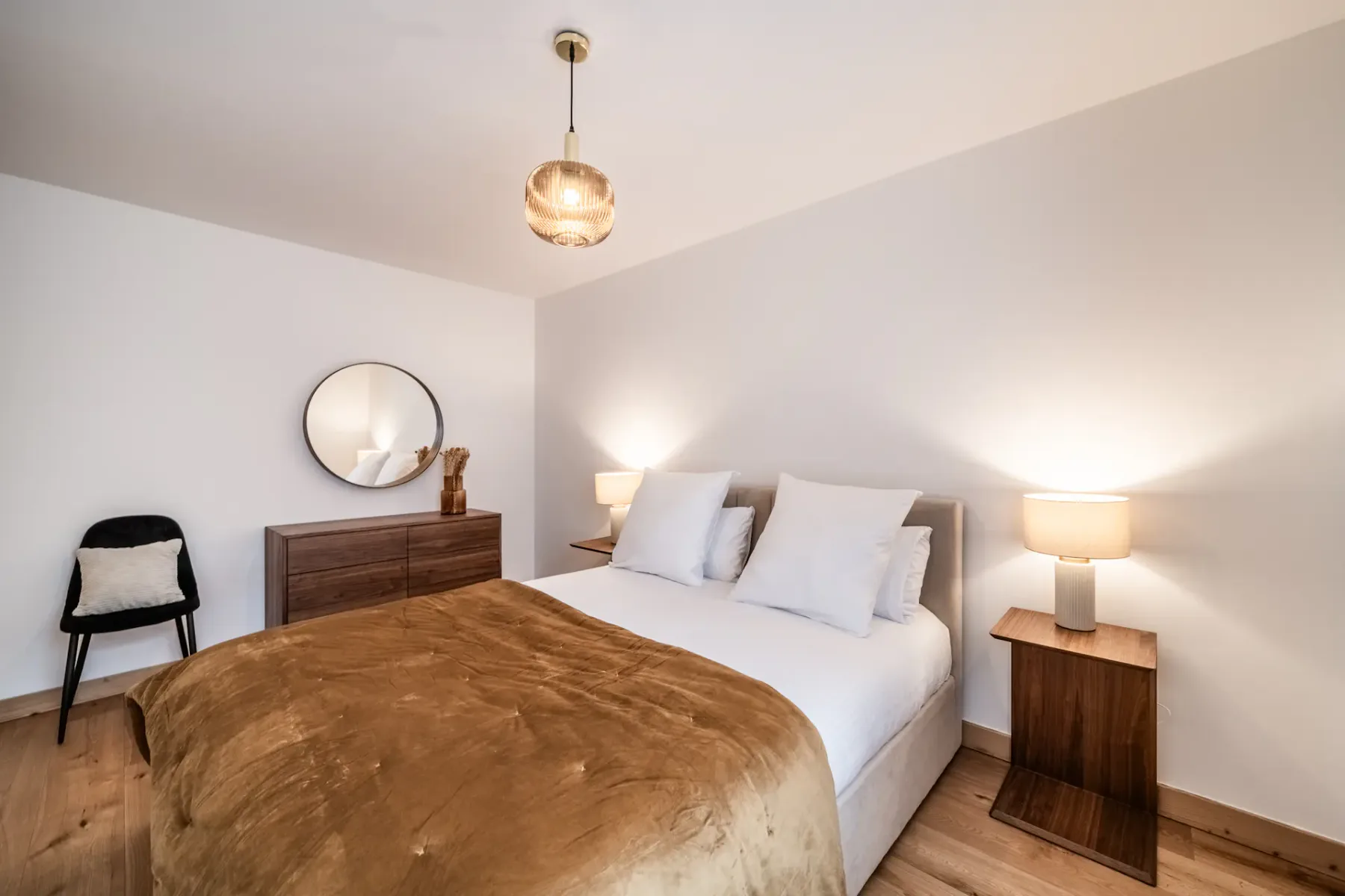 A well-lit bedroom with a double bed featuring white pillows and a brown duvet, two bedside tables with lamps, hardwood flooring, a pendant light, and a window with sheer curtains.