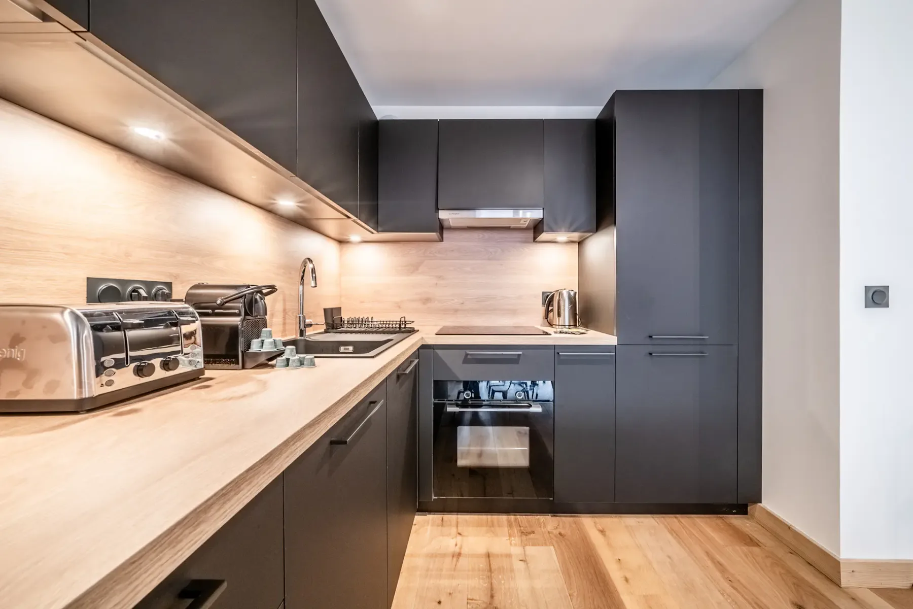 Modern kitchen with black cabinets, stainless steel appliances, and wooden countertops. Under-cabinet lighting illuminates the workspace. A dining table is partially visible on the left.