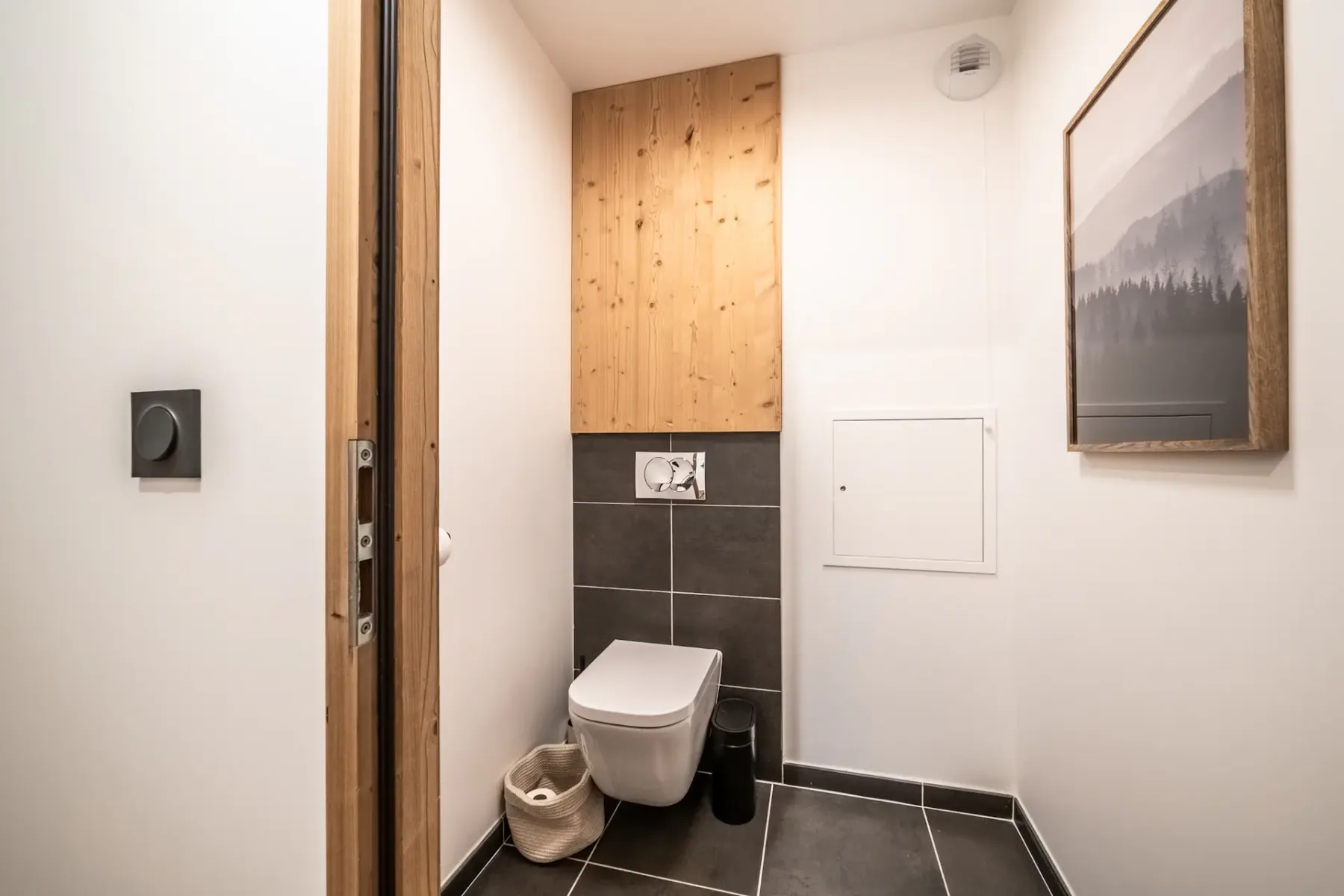 A modern bathroom with a wall-mounted toilet, wooden paneling above, and a framed nature picture on the right wall. A small trash bin and wicker basket are on the floor.