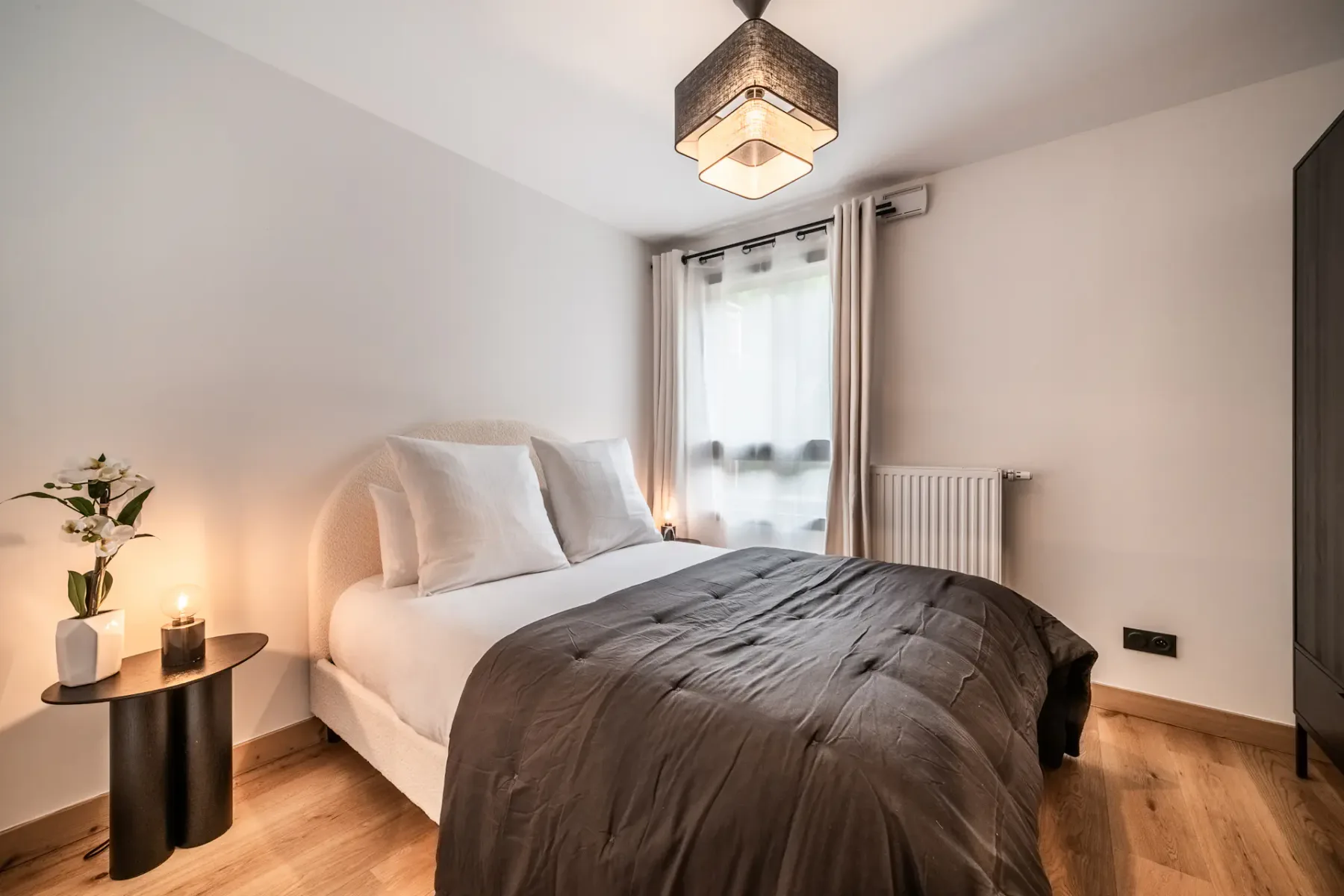 Modern bedroom with a neatly made bed, gray bedding, and white pillows. There's a side table with a lamp and a flower vase. A radiator and large window with sheer curtains are in the background.