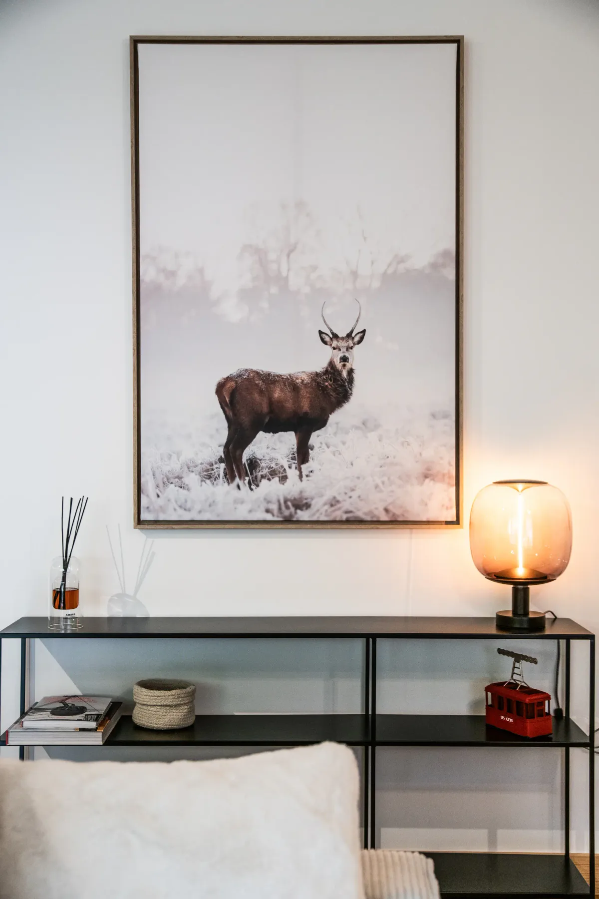Framed deer photo on a wall above a shelf with books, a vase, a reed diffuser, and a lamp emitting warm light.