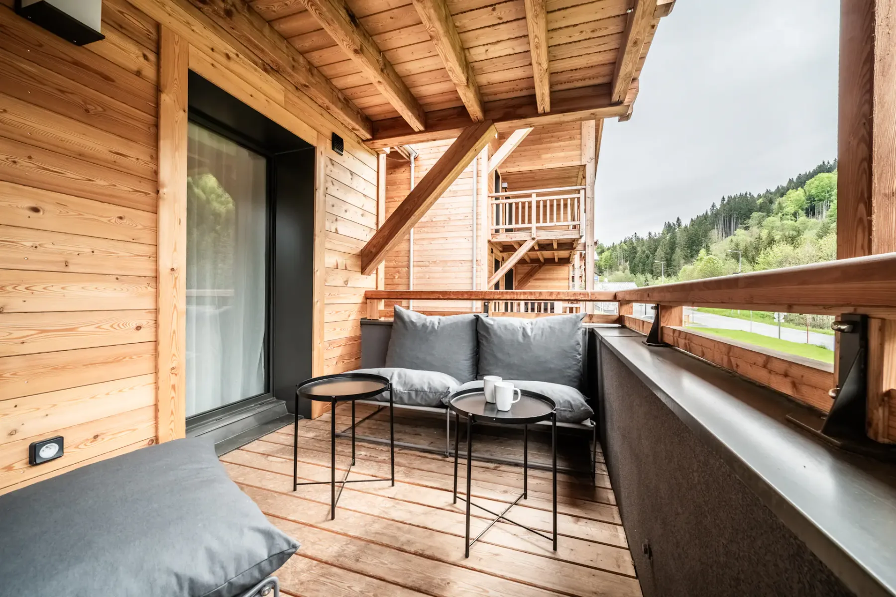Covered wooden terrace with cushioned seating and a view of green hills.