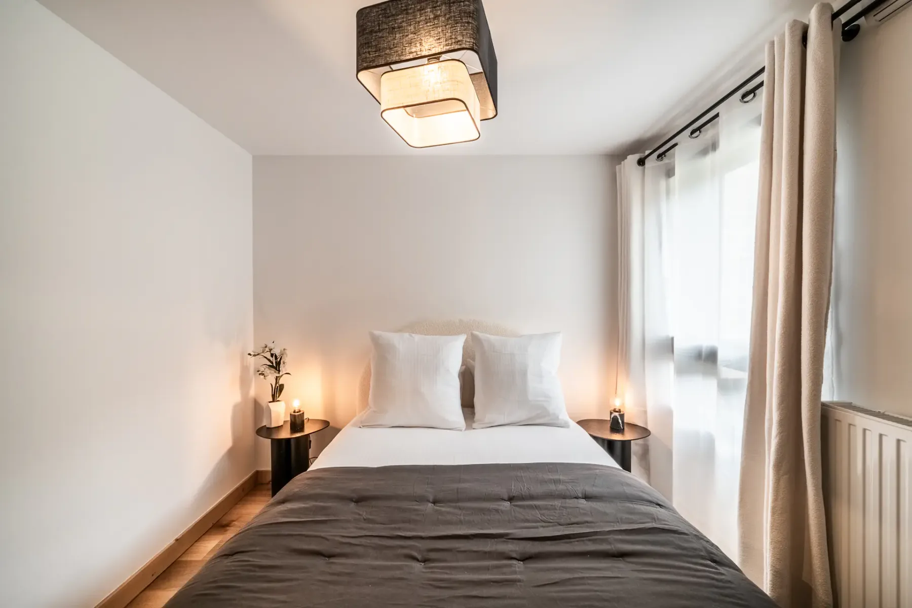 Modern bedroom with a neatly made bed, gray bedding, and white pillows. There's a side table with a lamp and a flower vase. A radiator and large window with sheer curtains are in the background.