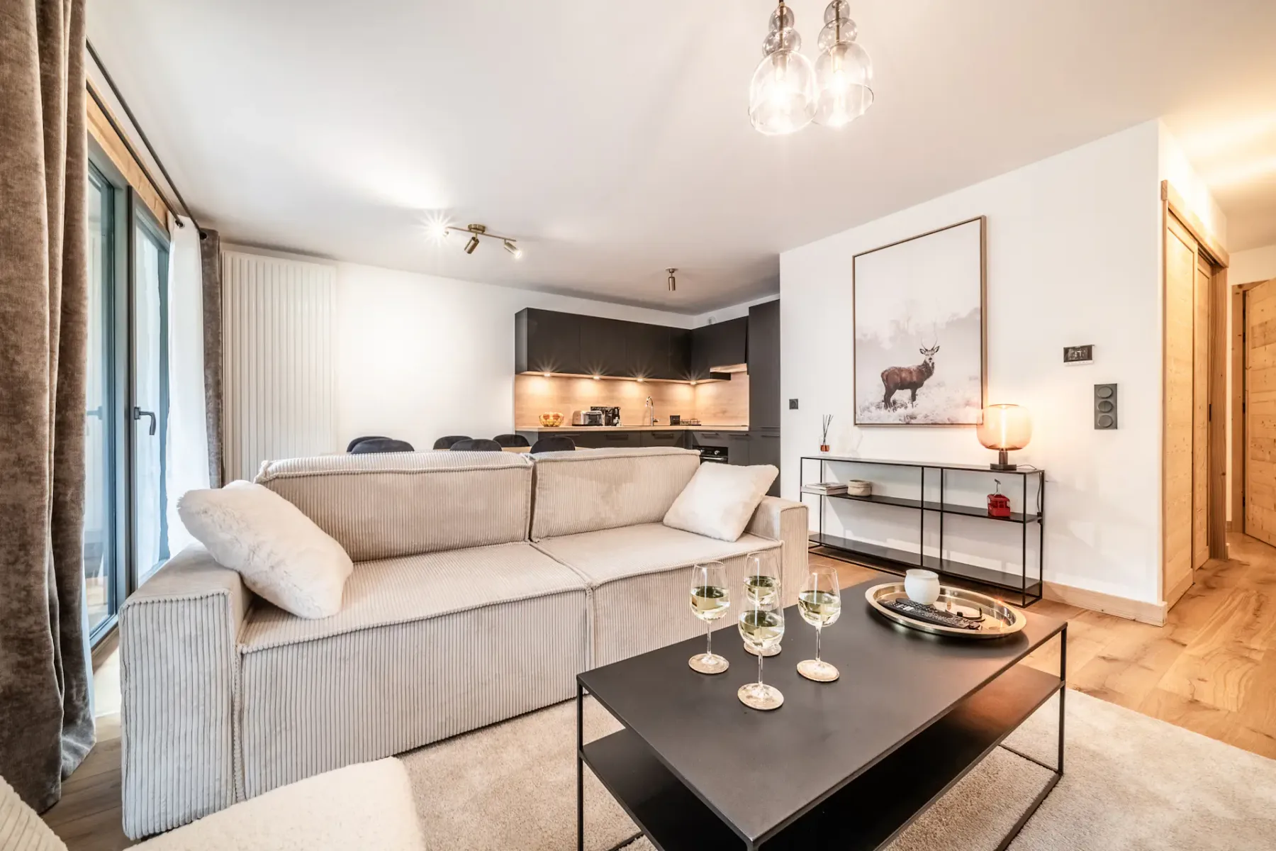 A bright living room with light-colored seating, a dark coffee table, and an open kitchen in the background.