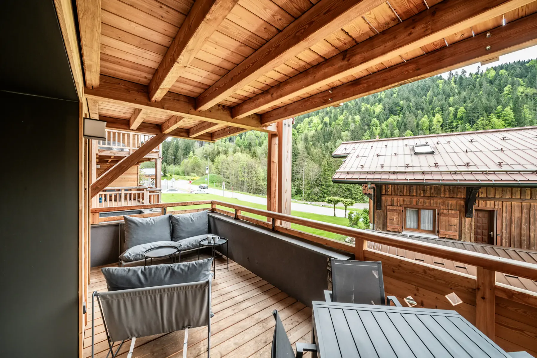 A covered wooden deck has outdoor seating and a view of a neighboring building and wooded hillside.