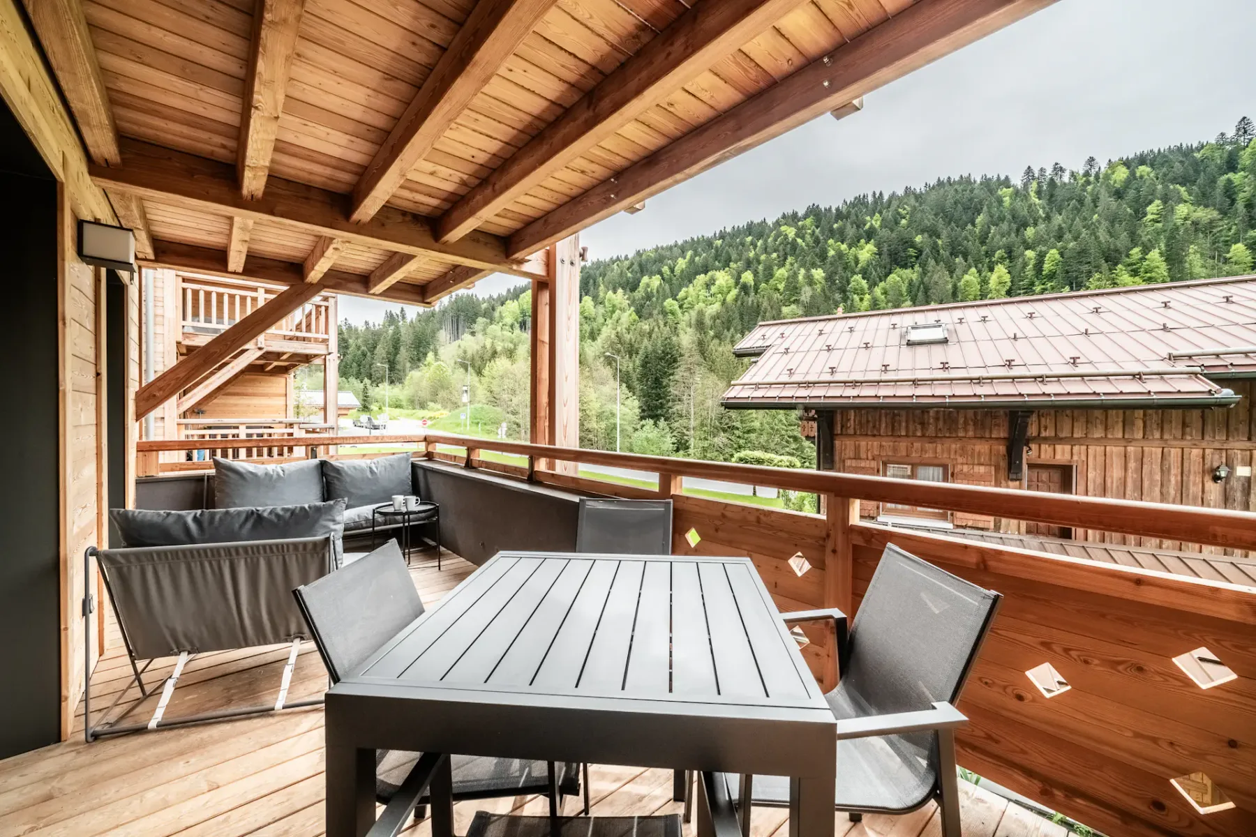 Outdoor deck or terrace with wooden roof, modern black table and chairs, and cushioned seating area. Overlooks wooded hillside and rustic buildings.