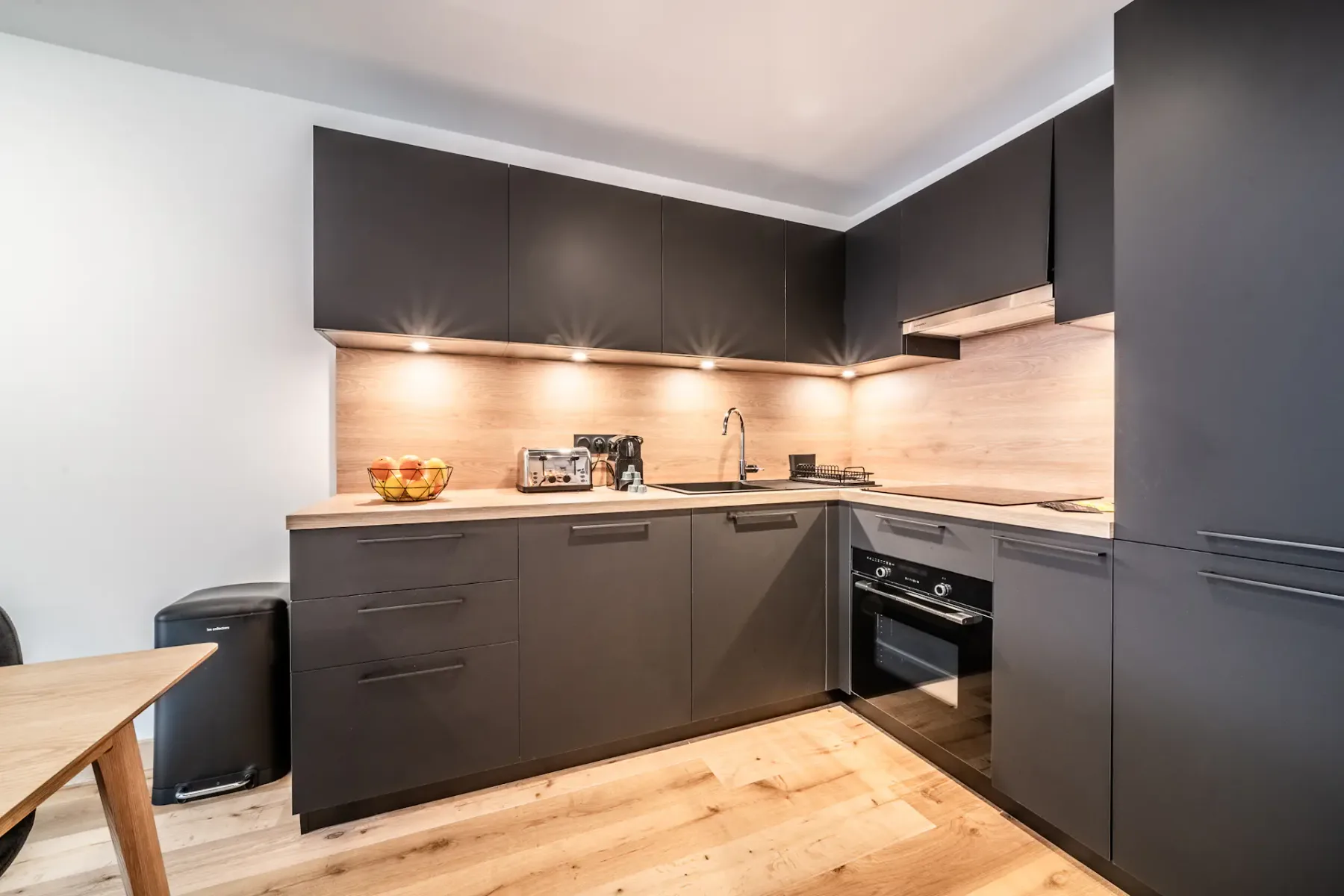 Modern kitchen with black cabinets, stainless steel appliances, and wooden countertops. Under-cabinet lighting illuminates the workspace. A dining table is partially visible on the left.
