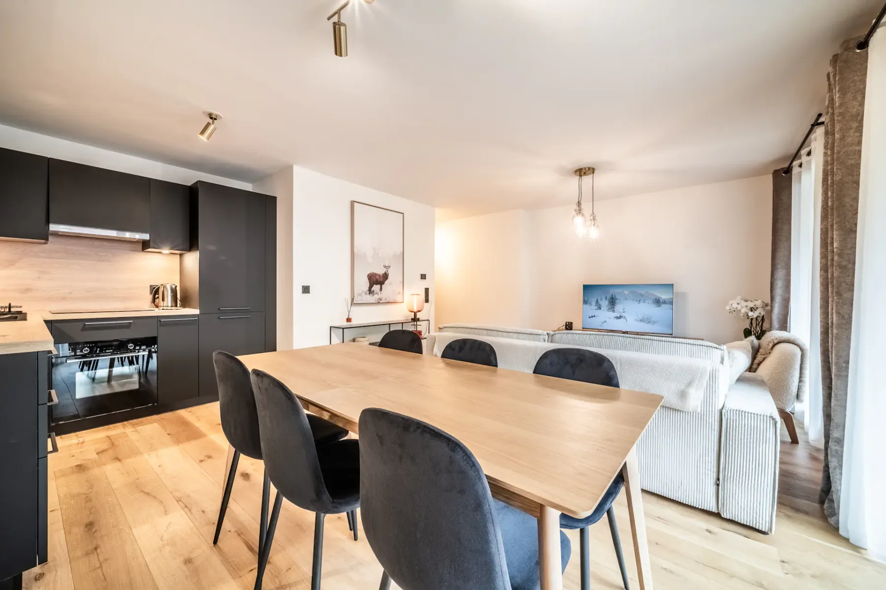 Modern living and dining area with a wooden table and black chairs. The kitchen has black cabinets. A TV is on, and there's a painting of a deer on the wall. The space is well-lit with natural light.