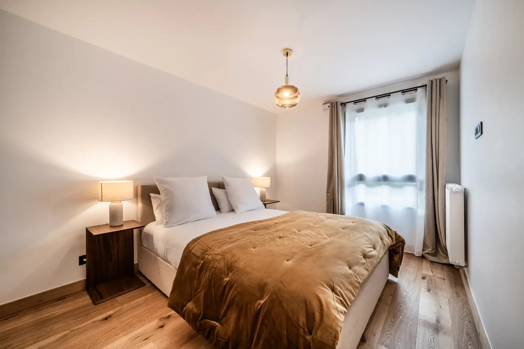 A well-lit bedroom with a double bed featuring white pillows and a brown duvet, two bedside tables with lamps, hardwood flooring, a pendant light, and a window with sheer curtains.