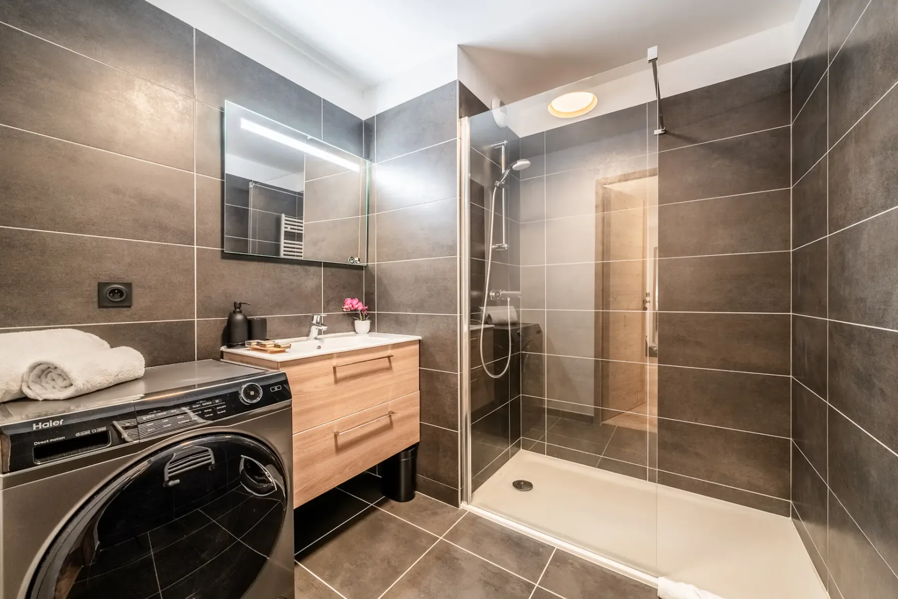 Modern bathroom with a glass-enclosed shower, floating wooden vanity with a sink and mirror, dark tiled walls and floor, a washing machine, and folded towels.
