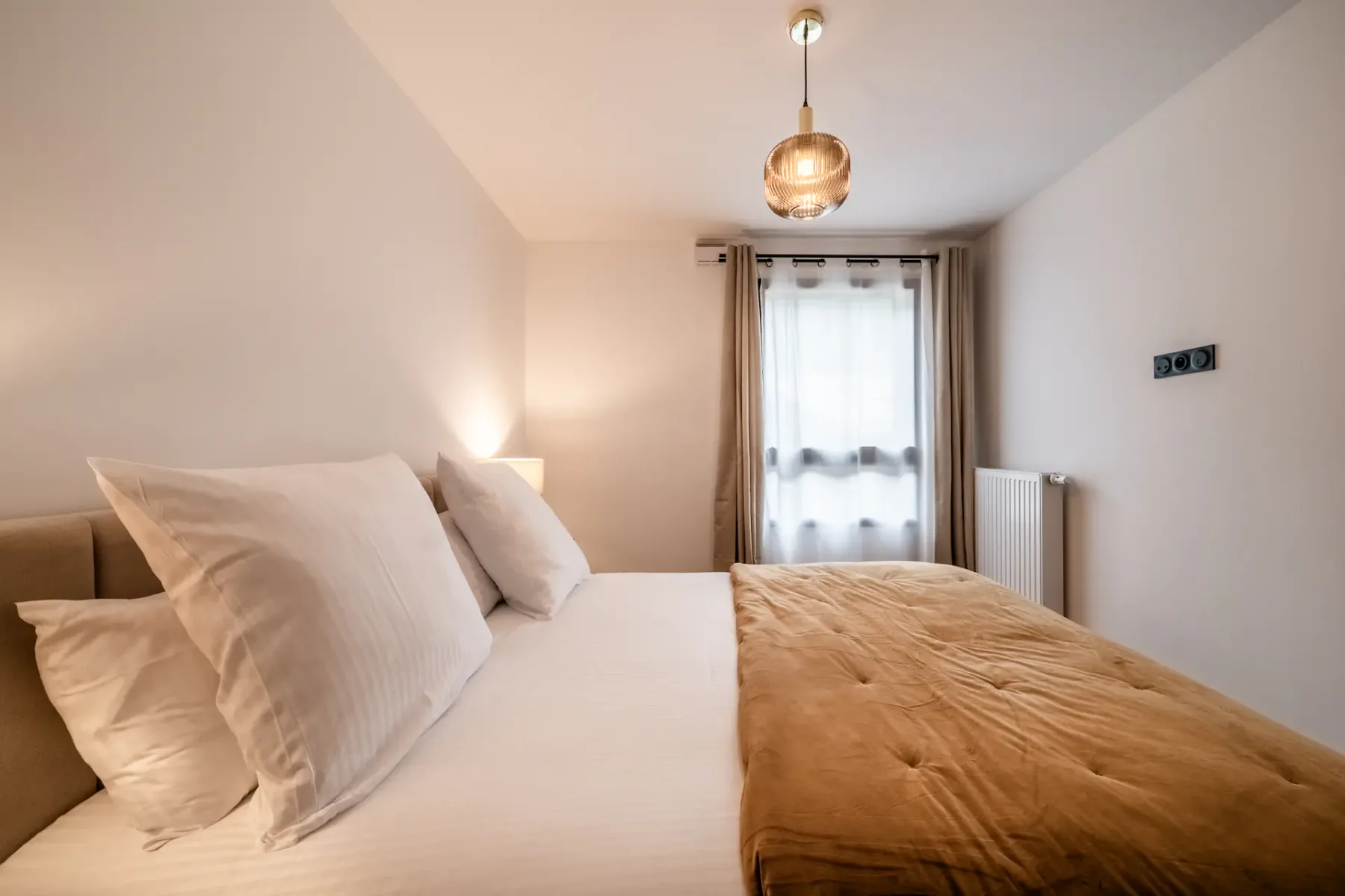 A bedroom with white bedding and a tan cover, with a curtained window at the end.