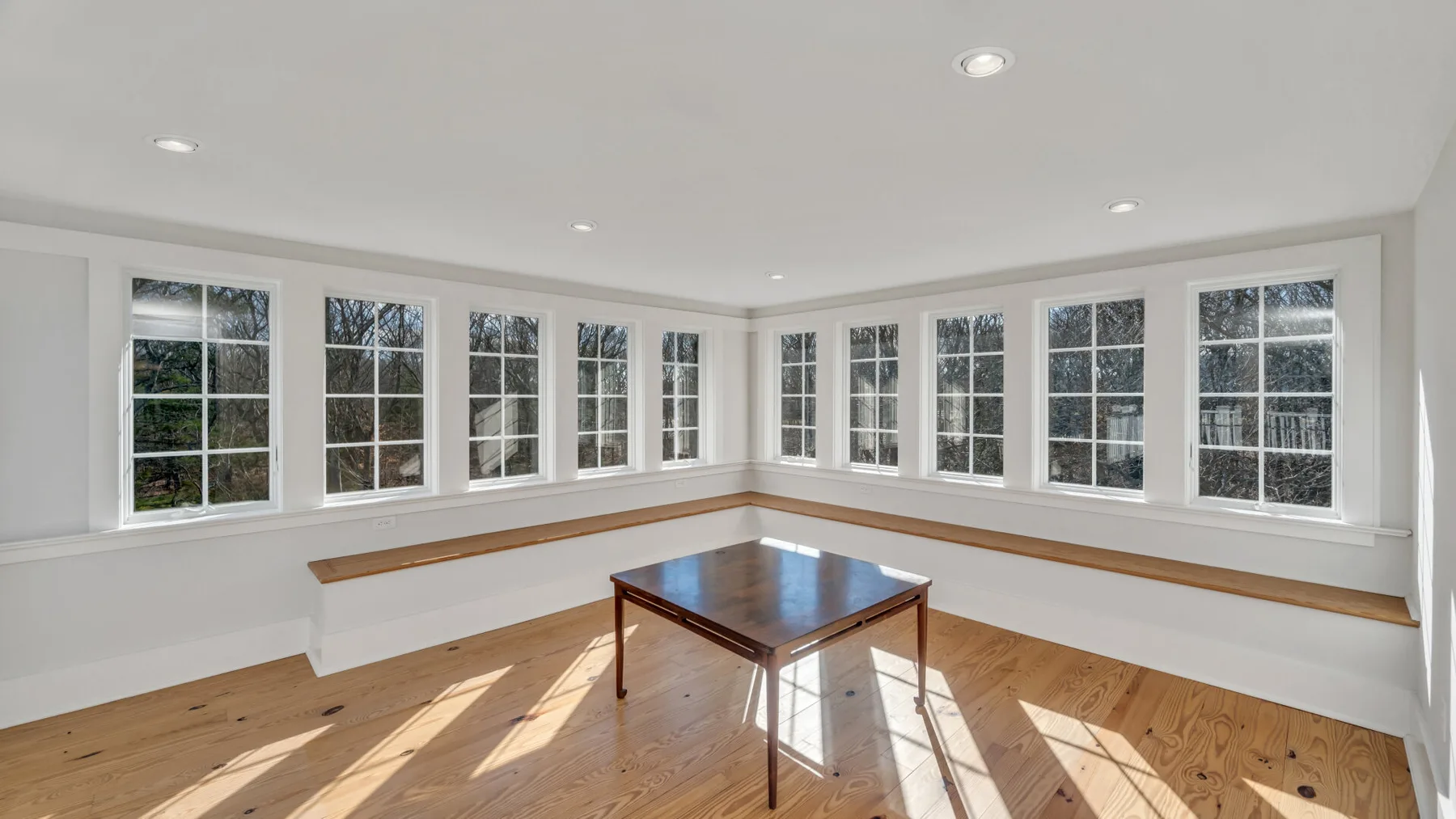 Una habitación luminosa con ventanas en dos paredes y una mesa de madera en el centro.