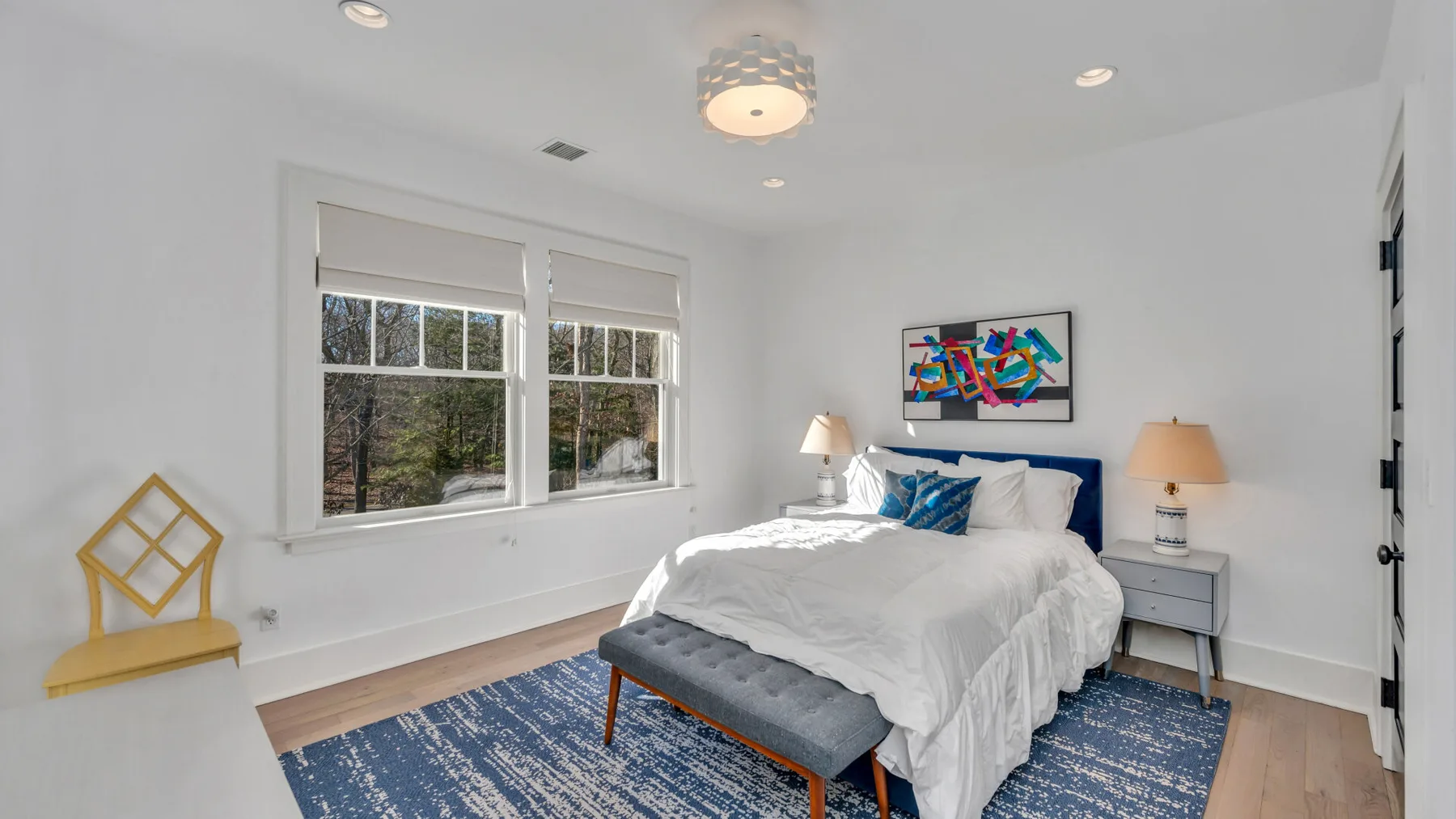Un dormitorio luminoso con una cama, dos ventanas y arte colorido sobre el cabecero.