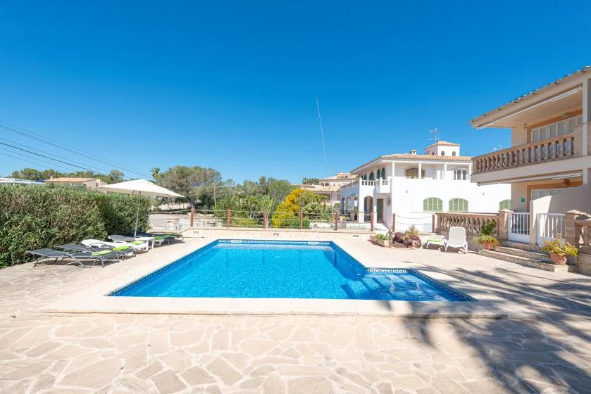 Una piscina exterior está junto a una terraza pavimentada y edificios blancos.