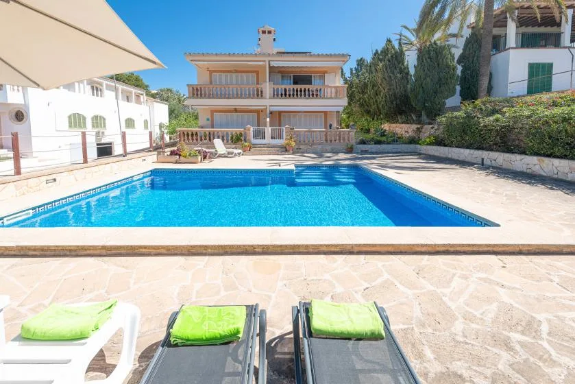 A swimming pool sits in front of a multi-story house, with lounge chairs nearby.