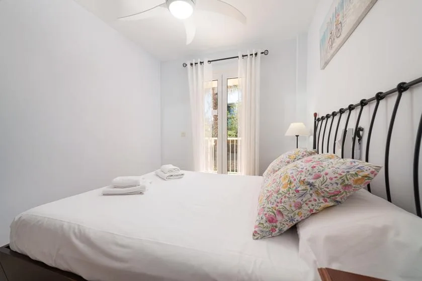Un dormitorio luminoso con una cama blanca, almohadas florales y una puerta acristalada con cortinas.