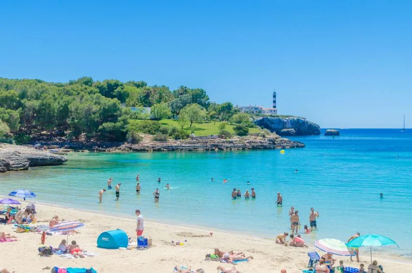 Una playa de arena junto a aguas azules, con bañistas y una costa arbolada.