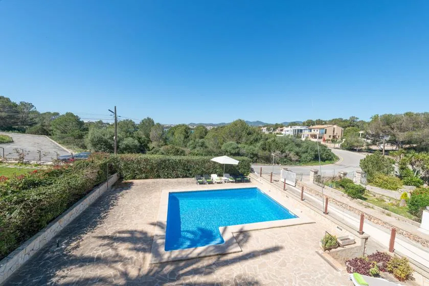 Una piscina está en una zona exterior soleada y pavimentada, rodeada de vegetación.