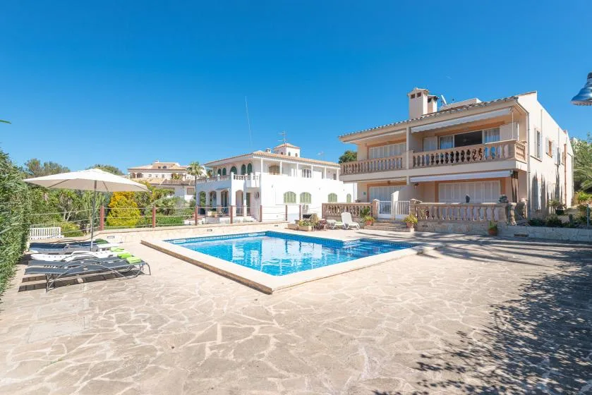 An outdoor swimming pool sits beside a large house on a sunny day.