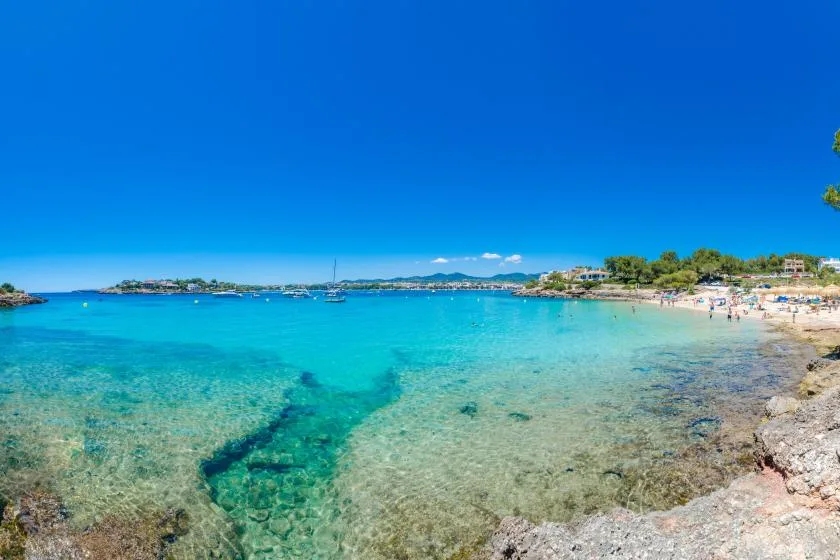 Una bahía costera de aguas turquesas, zonas rocosas poco profundas y una playa de arena.