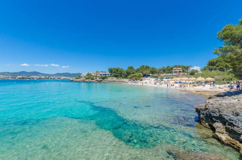 Una bahía costera turquesa se encuentra con una playa de arena y una orilla rocosa bajo un cielo azul despejado.