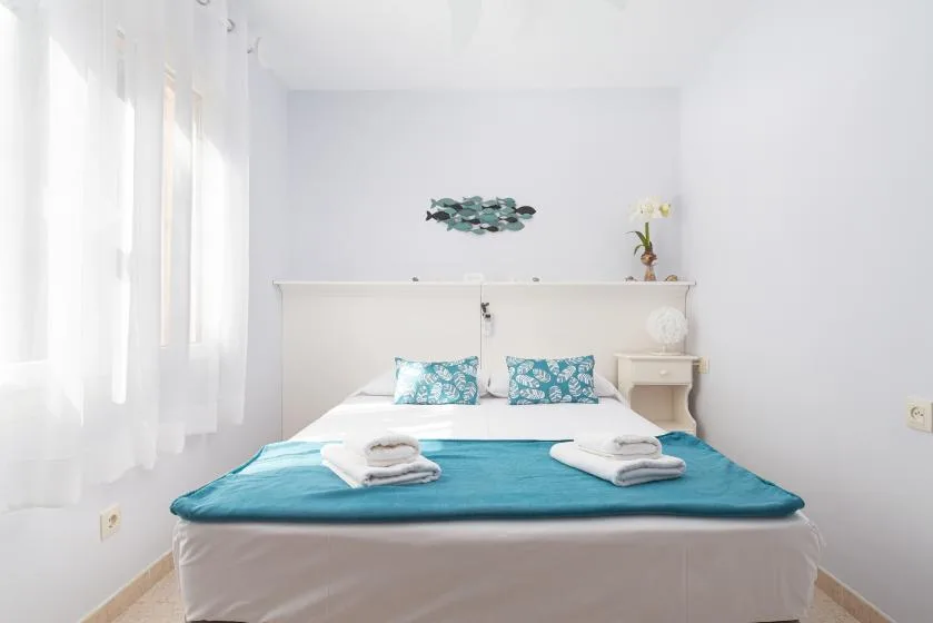 Un dormitorio luminoso con una cama cubierta por una manta turquesa y cortinas translúcidas.
