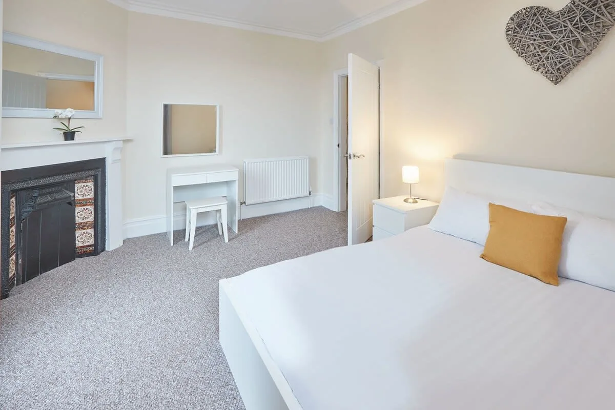 A bright bedroom with a white bed, bedside lamp, fireplace, and small desk.