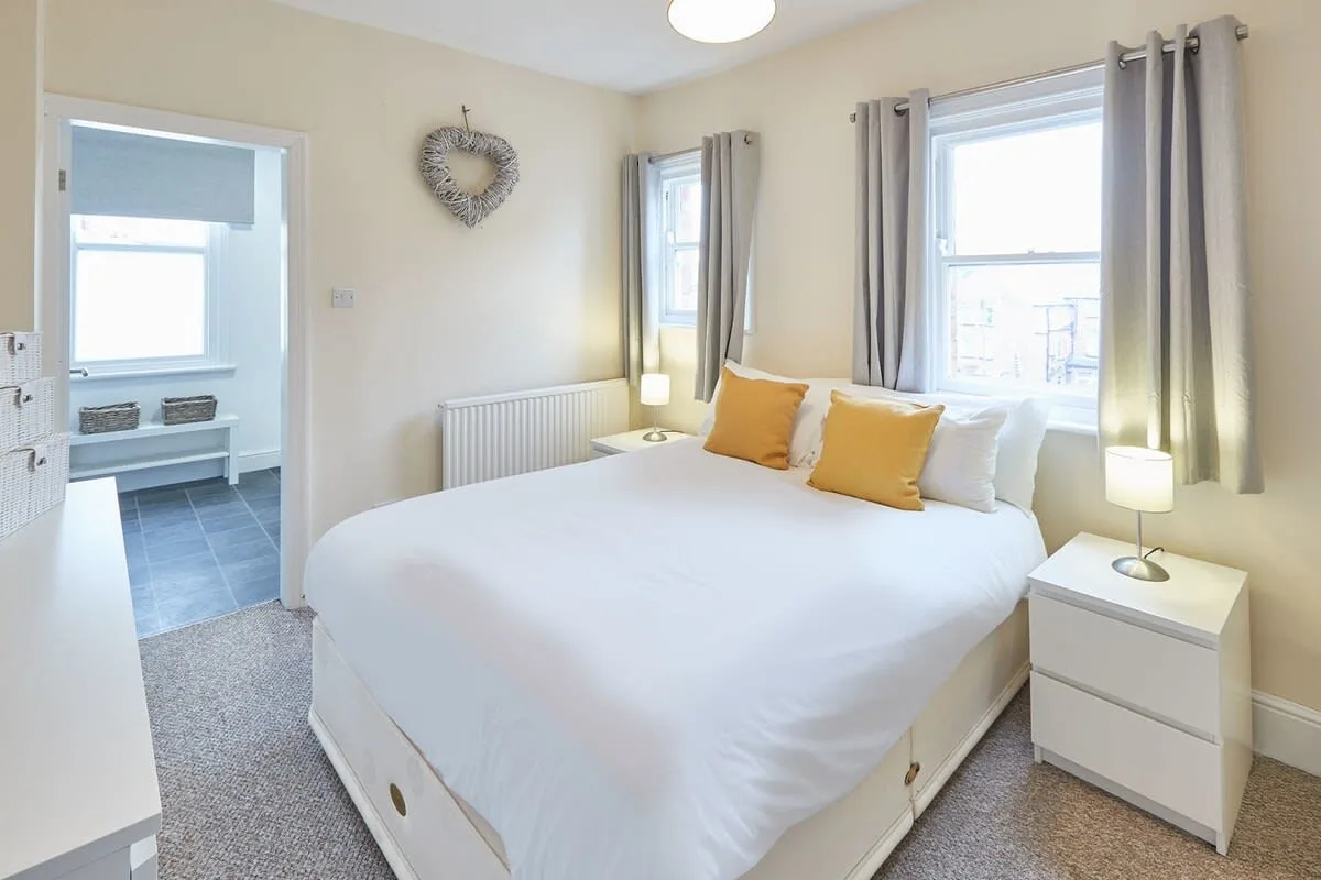 A bright bedroom with a neatly made bed and windows dressed with curtains.