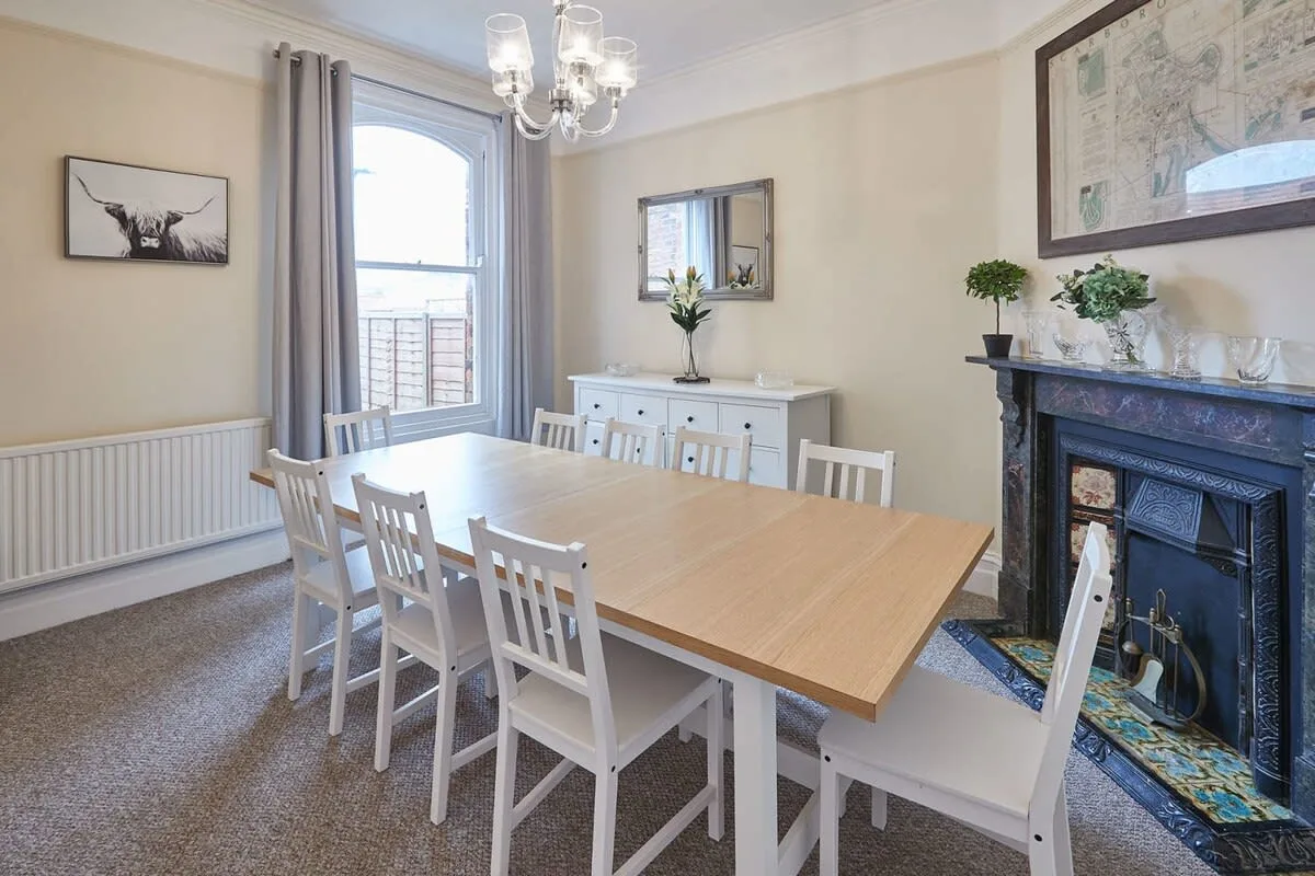 A dining room with a long wooden table and white chairs beside a fireplace.