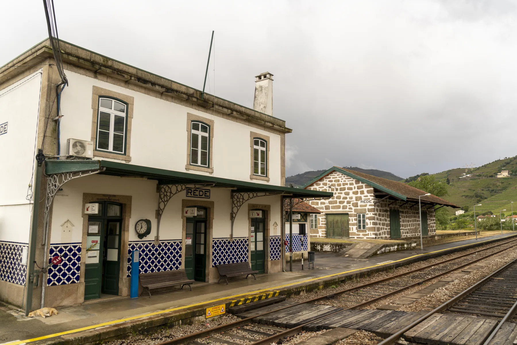 Un edificio de estación de tren junto a las vías, con una construcción de piedra cercana.